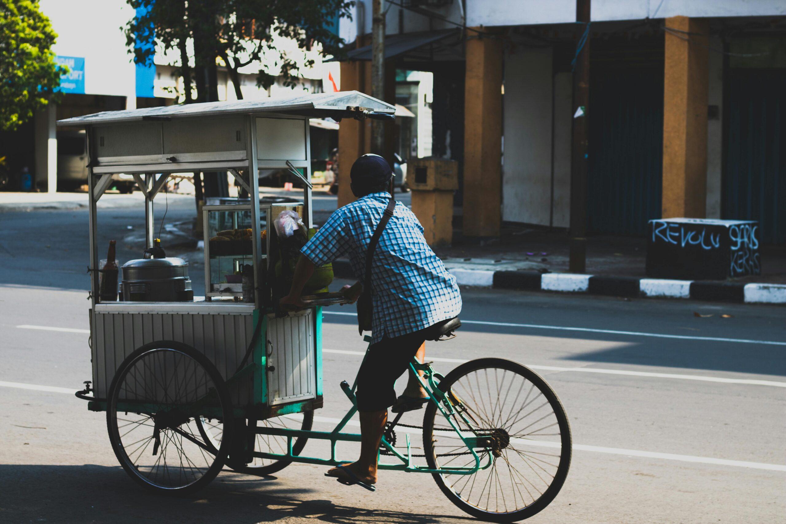 An adult male vendor rides a bicycle food cart through a city street on a sunny day